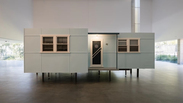 A light blue fibro home with two windows and a door on stilts 