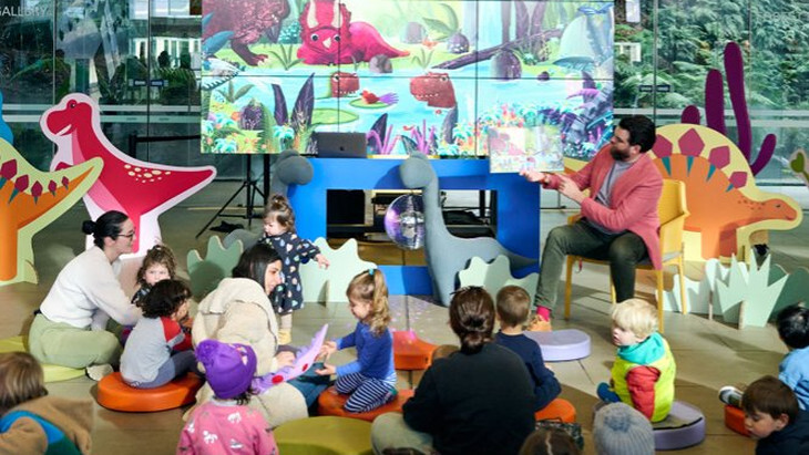 Children and parents listening to a story book beside large dinosaur cutouts at Melbourne Museum