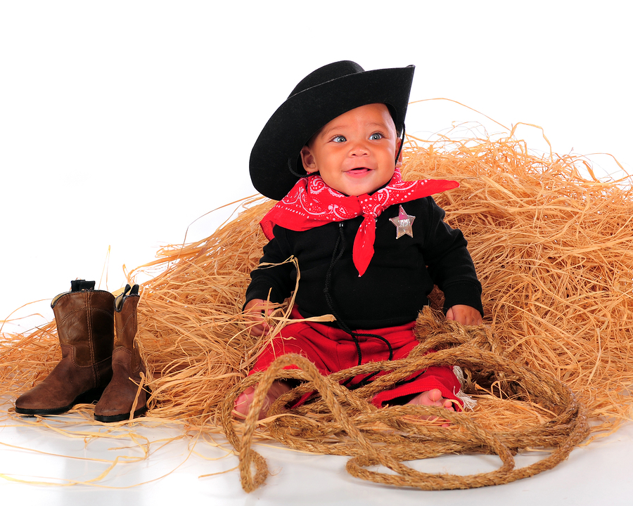 A happy biracial baby dressed as a cowboy sitting in a pile of hay. Isolated on white.